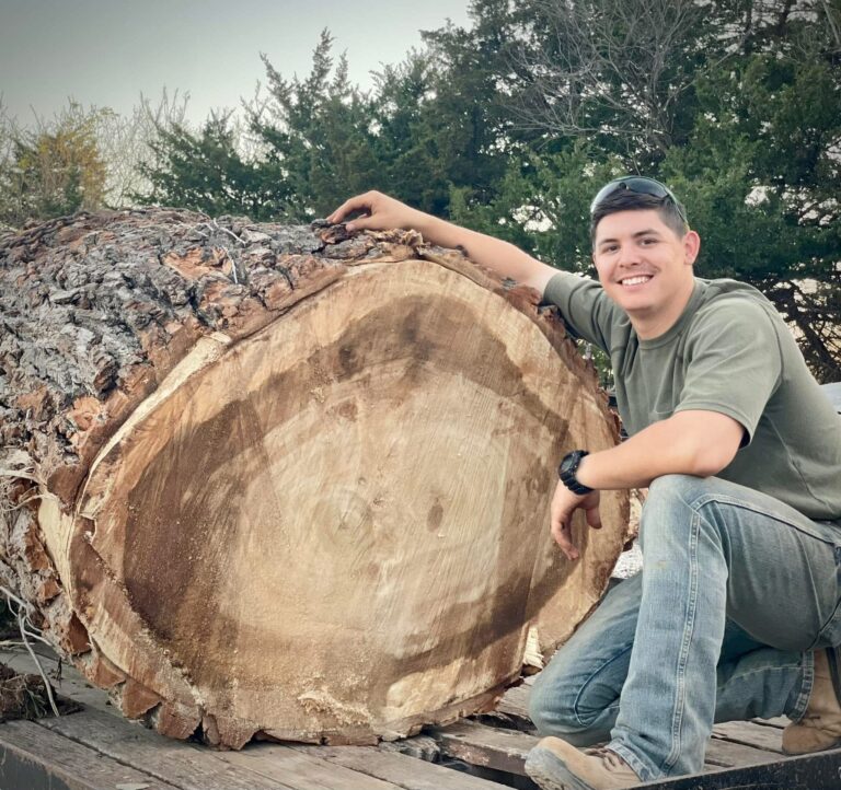 A smiling person in a green shirt and jeans kneels beside a massive tree trunk lying on a flatbed trailer, with trees and greenery in the background. - Tree Bid Pro